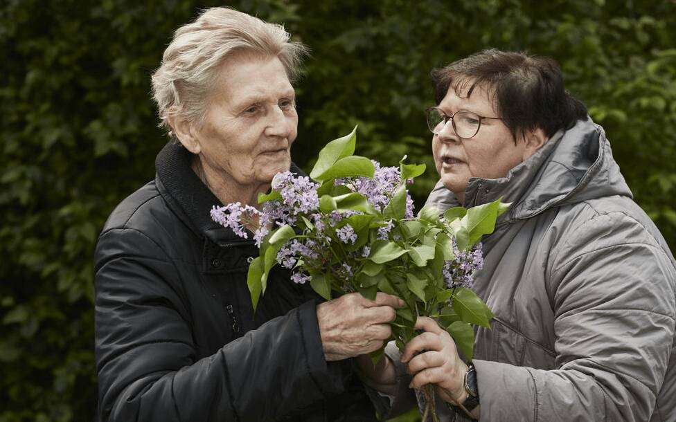 Eine alte Dame und ihre Begleiterin stehen im Garten mit einem Strauß Flieder im Arm.