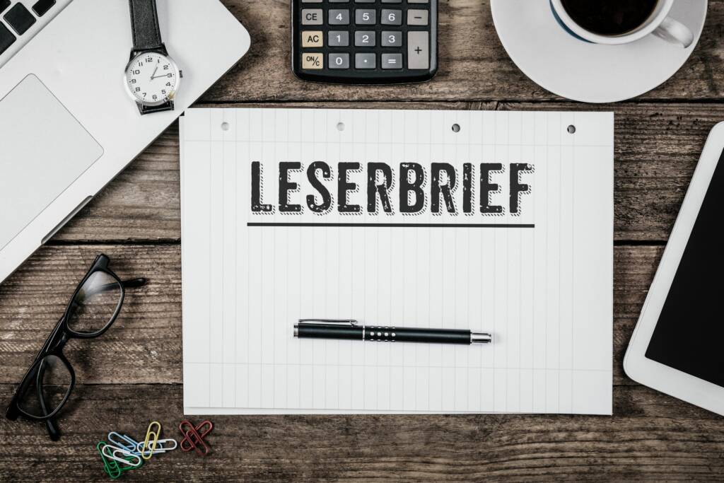Der Bildsausschnit zeigt einen Schreibtisch von oben. In der mitte ein Notizblock im Querformat mit dem Schriftzug "Leserbrief" und einem Kugelschreiber darunter. Drumherum liegen weitere Office Gadgets wie Laptop oder Tablet.
