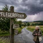 Wegweiser mit der Aufschrift "Pflegereform" an einer Landstraße. Im Hintergrund zieht ein Gewitter auf